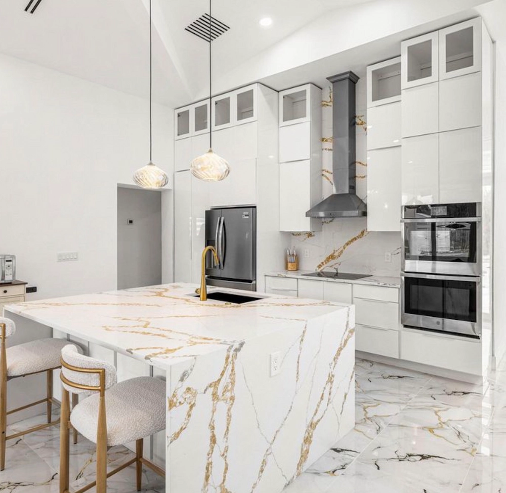 Luxury modern kitchen with white and gold veined marble island and floor, high-gloss white cabinetry, gold faucet, stainless steel appliances, double wall oven, and two pendant globe lights.